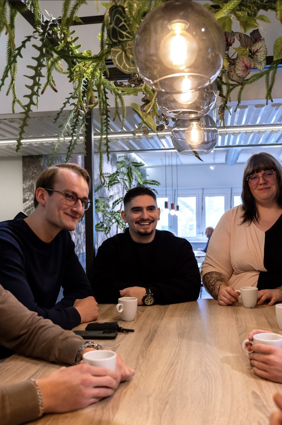 Drie Tolkie-collega's lachend aan een houten tafel met koffie in het kantoor